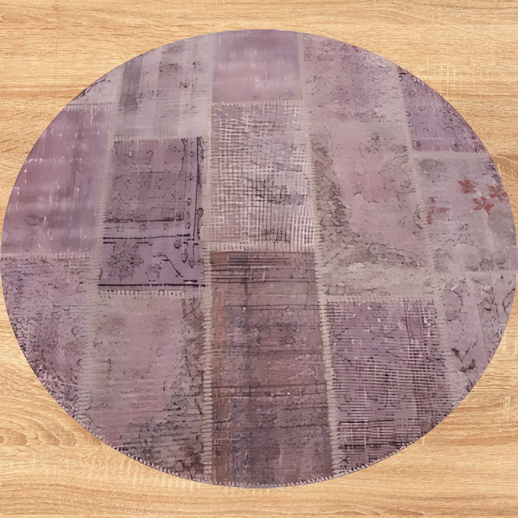 10_10 A round rug with a distressed, patchwork-style pattern in shades of mauve, lavender, and dusty rose. The rug is displayed on a light wood floor, showing its soft, muted tones.