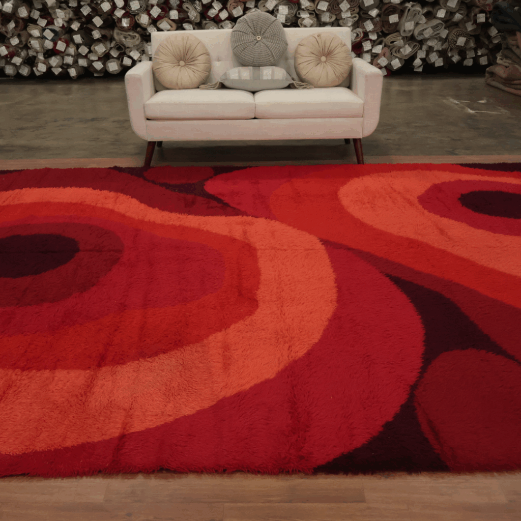 A close-up view of a retro-inspired shag rug in rich red and orange tones with curved, layered patterns. A cream couch with round pillows and rows of rolled carpets is visible in the background.