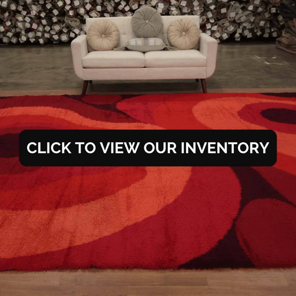A modern beige couch with round pillows sits behind a bold red and orange shag rug featuring wavy, abstract patterns. Rolls of carpet line the background, and a black overlay in the center reads, “Click to view our inventory.”