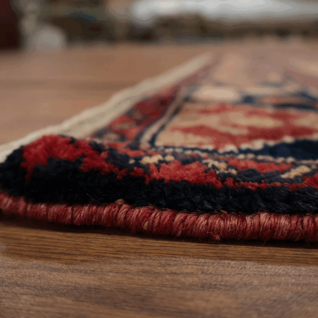 A detailed side view of a traditional woven rug with red and navy edging on a hardwood floor, showing the texture and weave of the material up close.