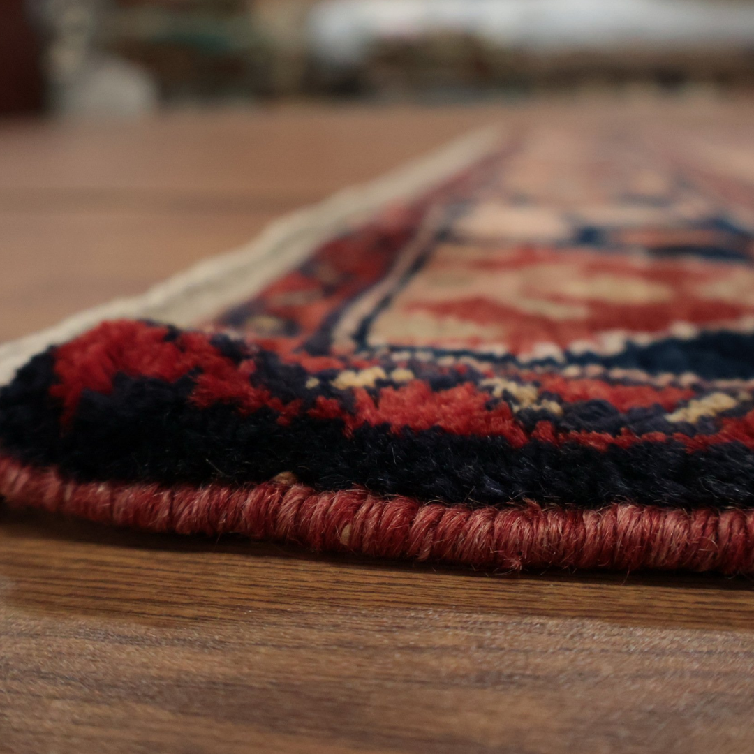 A detailed side view of a traditional woven rug with red and navy edging on a hardwood floor, showing the texture and weave of the material up close.