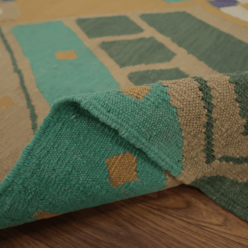 A close-up of a woven rug in teal and beige tones, featuring geometric block patterns. One corner is folded over to highlight the texture and thickness of the weave.