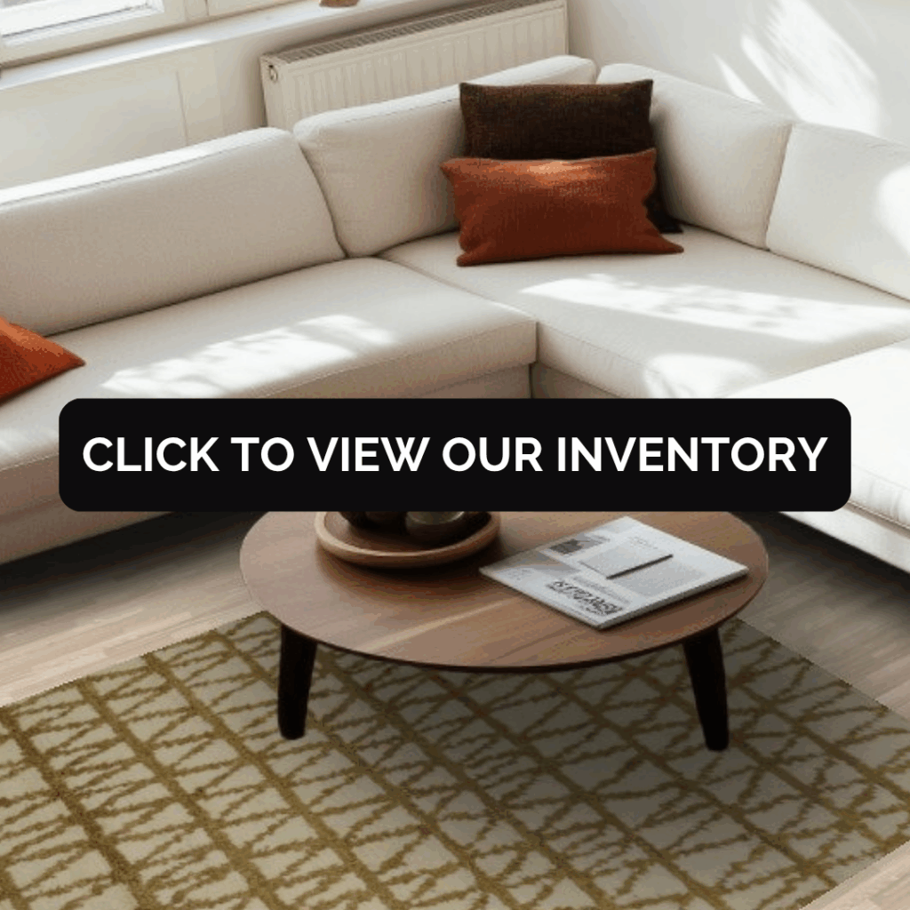 A sunlit room with a white sectional sofa and orange accent pillows surrounding a round wooden coffee table. A beige rug with a crisscross pattern anchors the space.