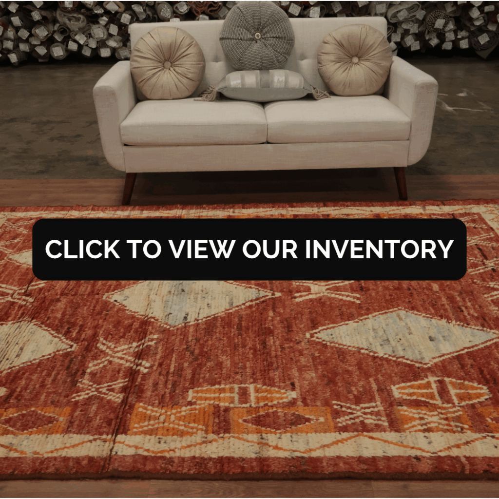 A neutral-toned loveseat with round accent pillows sits on a rich red-orange rug featuring cream and gold geometric patterns. Rolls of carpet are stacked in the background of a showroom. Text overlay reads “Click to View Our Inventory.”