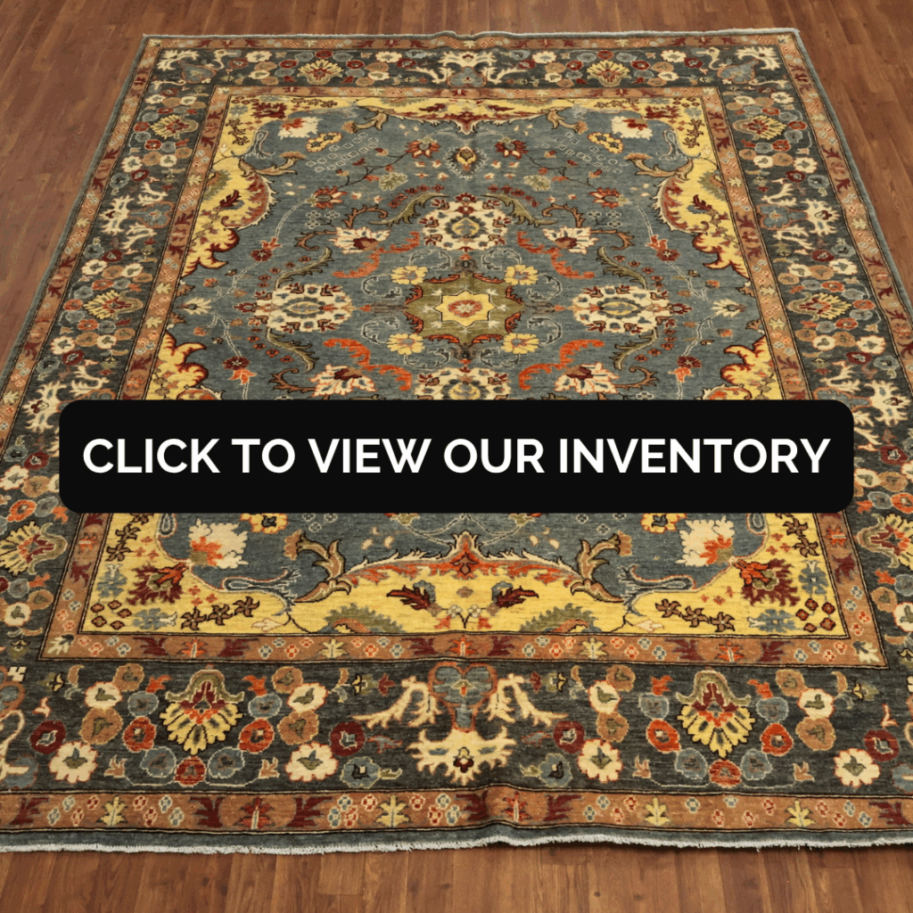 An ornate traditional rug with an intricate floral and scroll pattern in blue, gold, red, and cream tones is displayed on a wooden floor. Text overlay reads “Click to View Our Inventory.”
