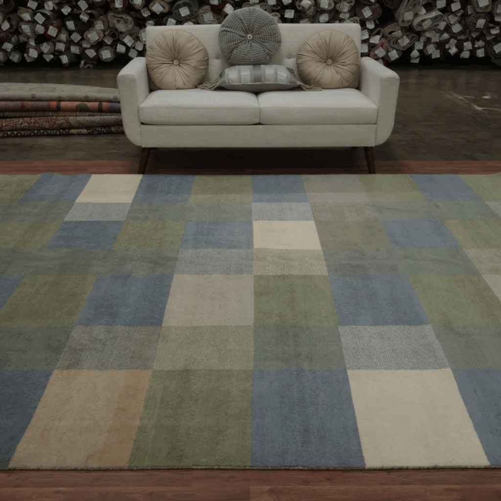 A modern rug in muted blue, green, beige, and cream squares lies in front of a cream sofa with round pillows, with rolled carpets stacked in the background.