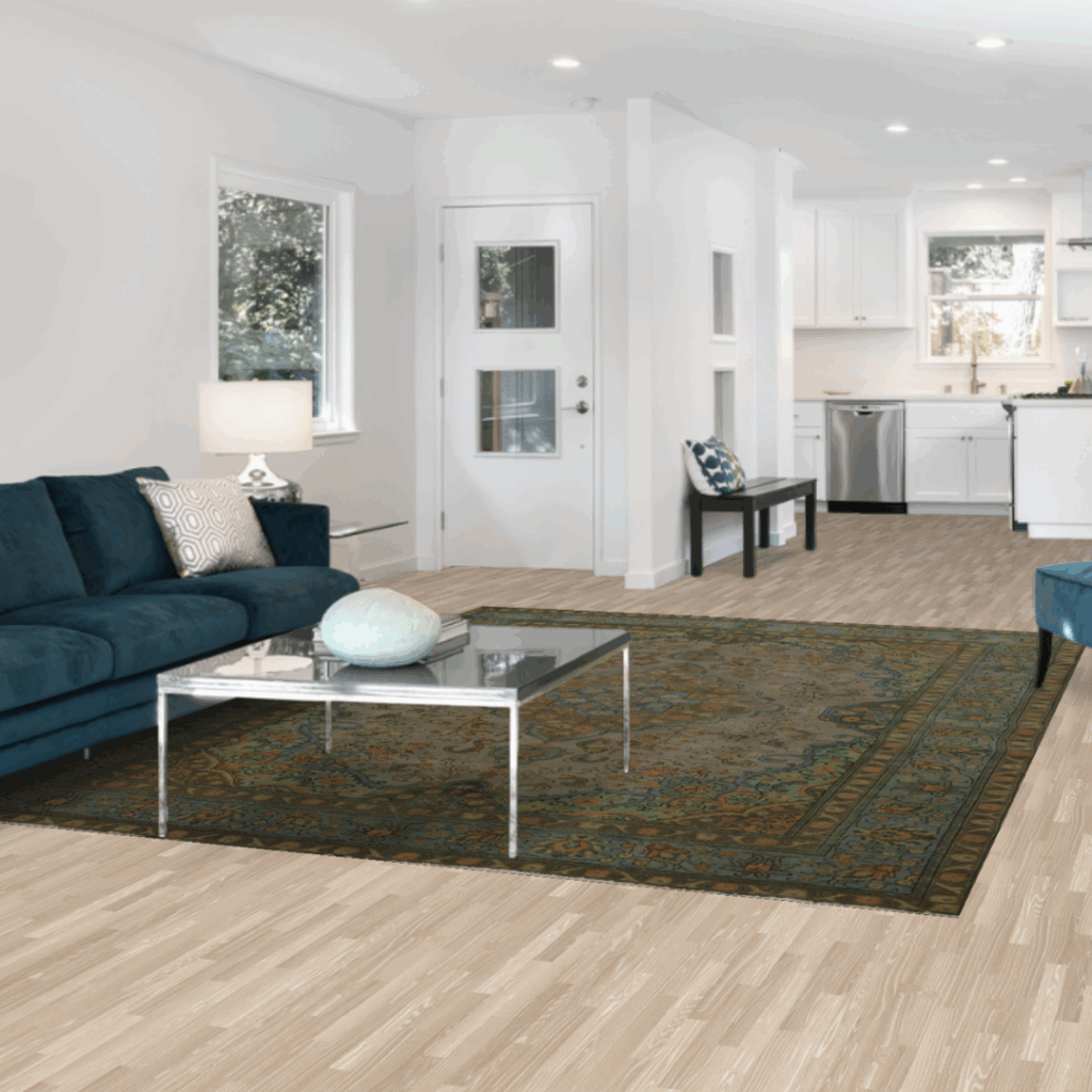 A bright, modern living room with white walls, teal seating, and a large green patterned rug under a glass coffee table, opening into a white kitchen.